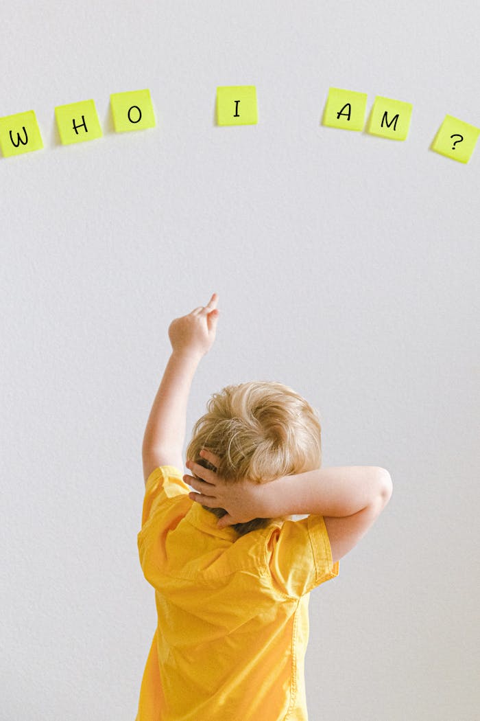Blond child in yellow shirt pointing at words 'WHO I AM?' on sticky notes on a wall.