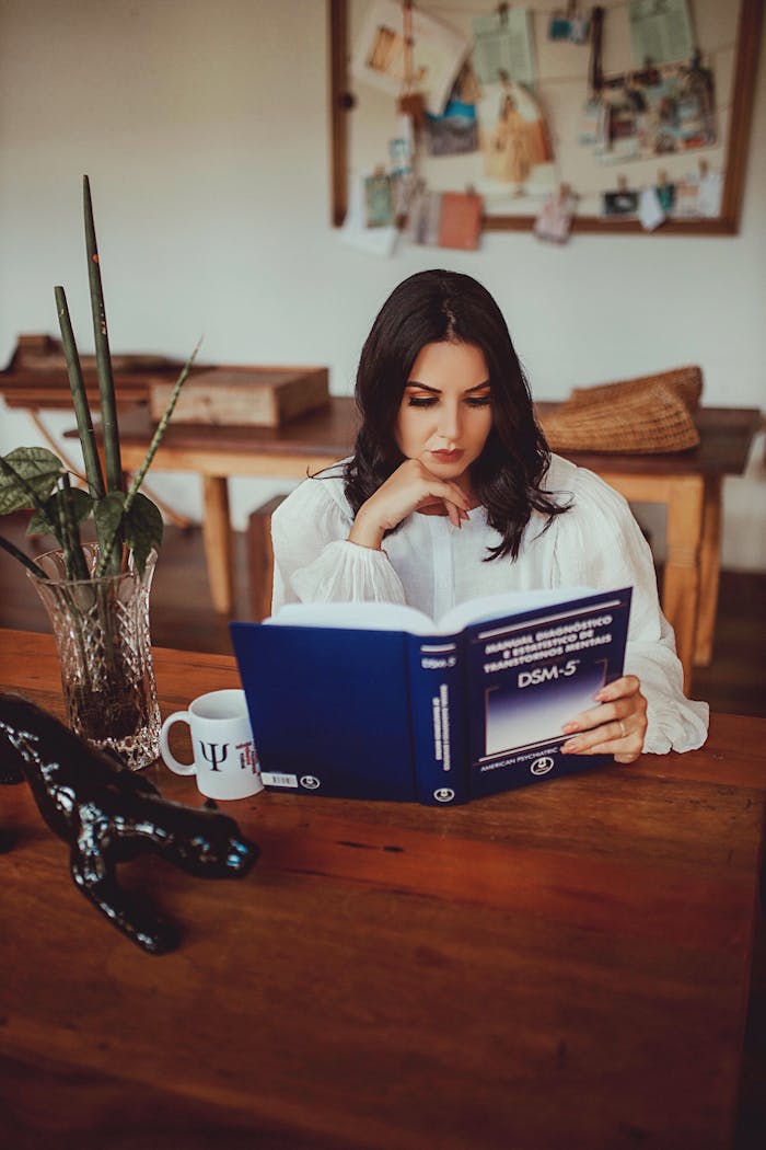 A young woman engrossed in reading the DSM-5 book at a wooden desk with a coffee mug in a cozy study environment.